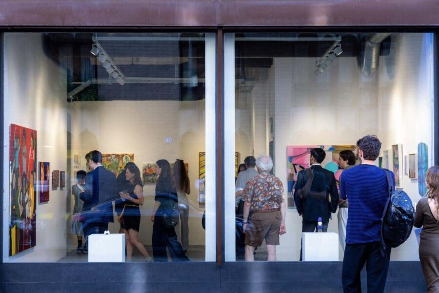 People viewing colorful paintings inside a brightly lit art gallery seen through a storefront window.