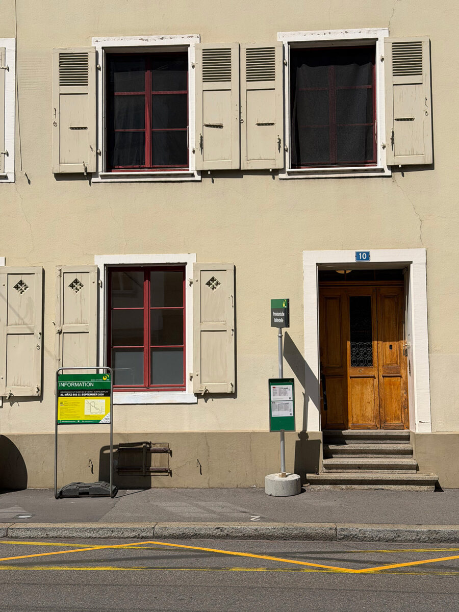 Beige building facade with red-framed windows, shutters, a wooden door, and information signs.