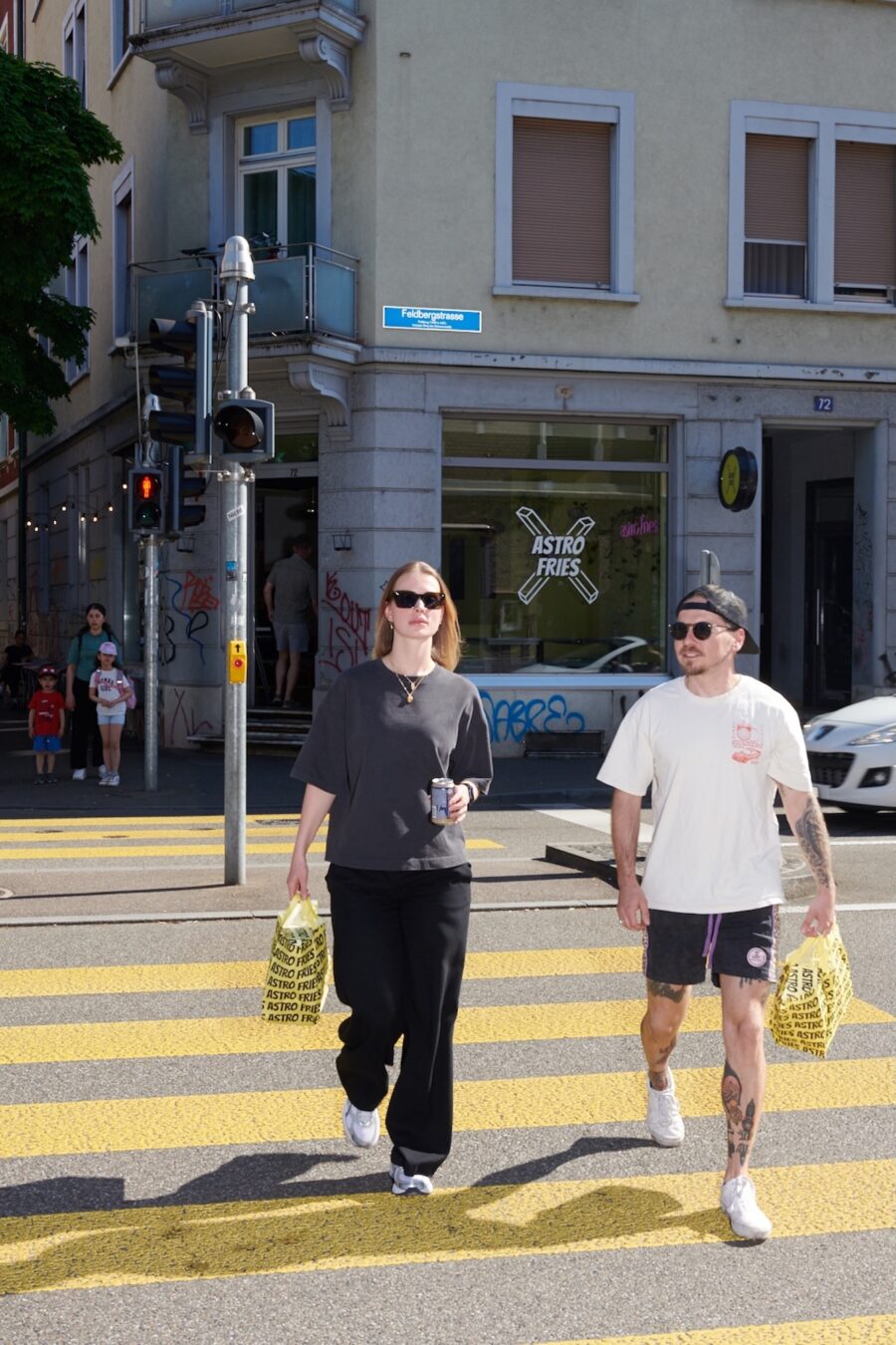 A man and woman carry Astro Fries bags while crossing a sunny city street.