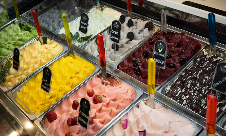 Variety of colorful gelato flavors in a display case with toppings and serving spatulas.