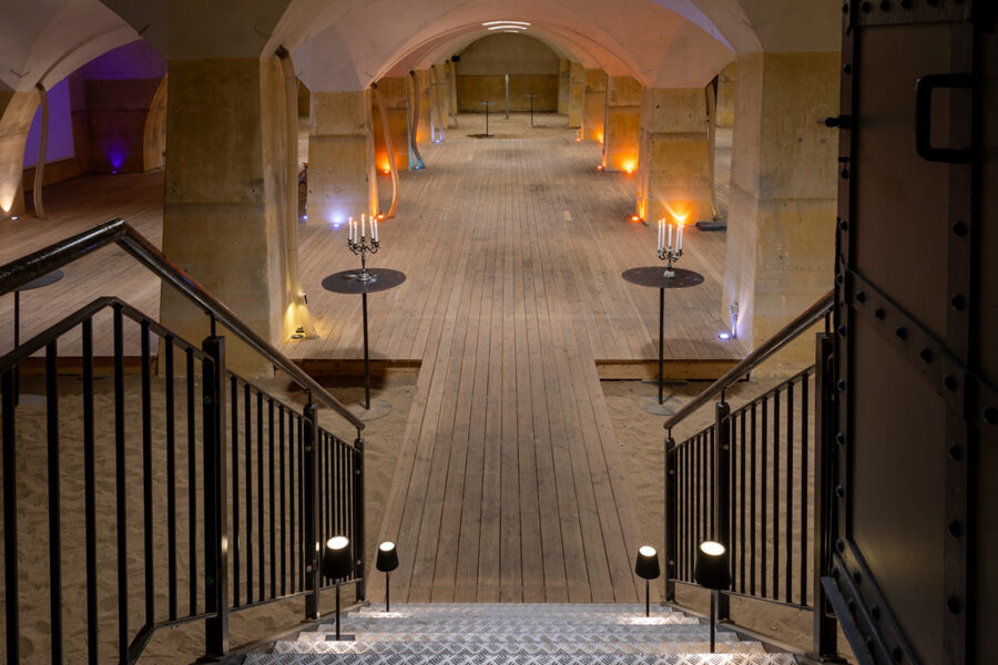 View from a staircase looking down into a vaulted stone cellar with a wooden walkway and candles.
