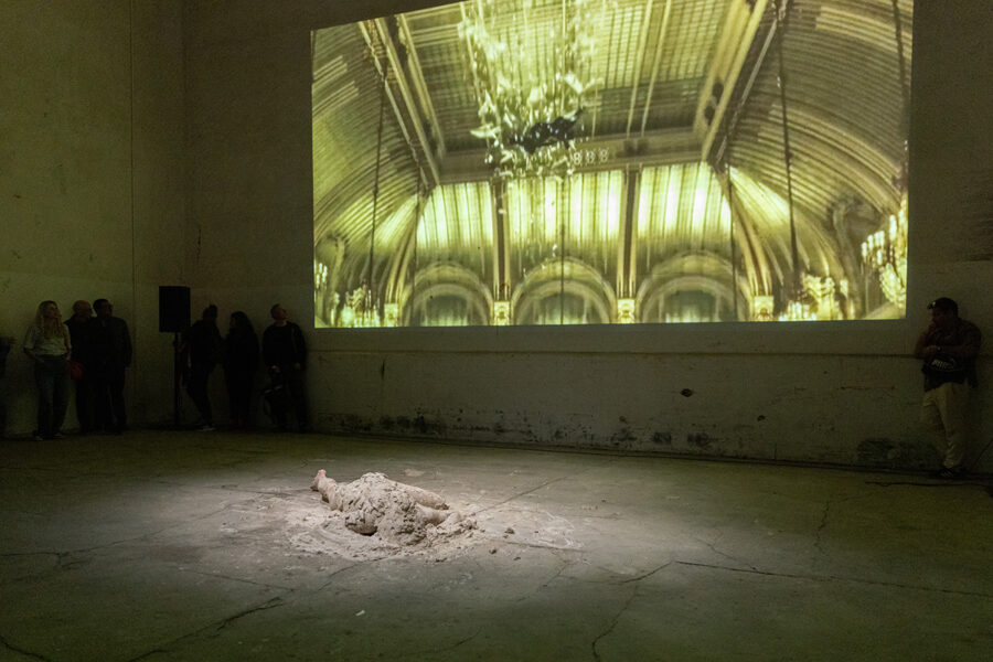 An art installation with a projection of an ornate hall and a sand sculpture on the floor.