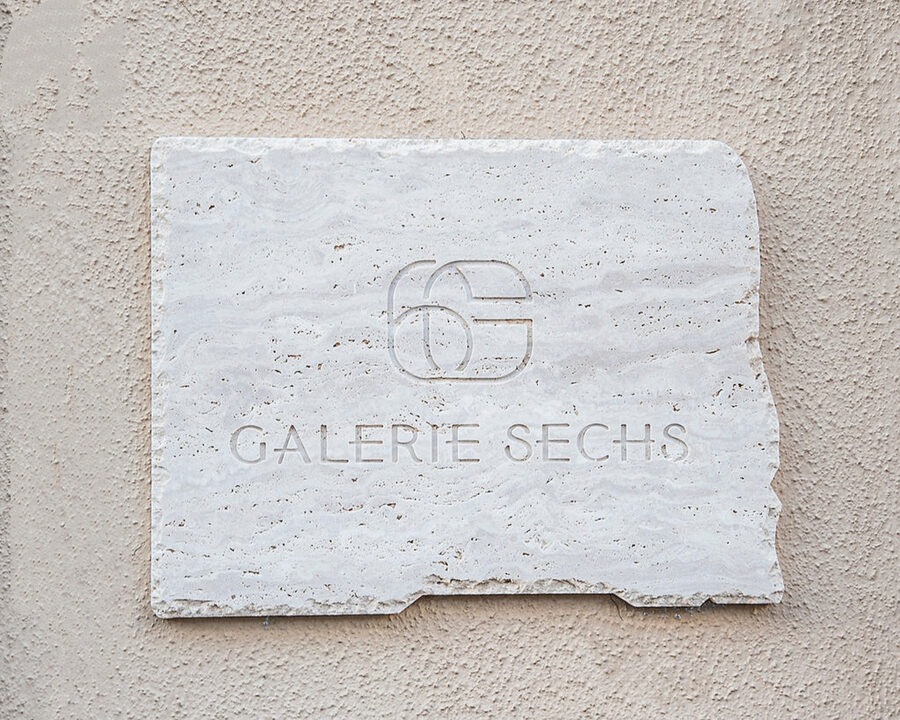 A white stone plaque on a textured wall engraved with a logo and the text GALERIE SECHS.