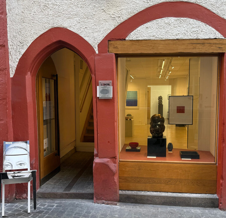 Exterior of an art gallery with a red stone archway and a display window showing sculptures and art.