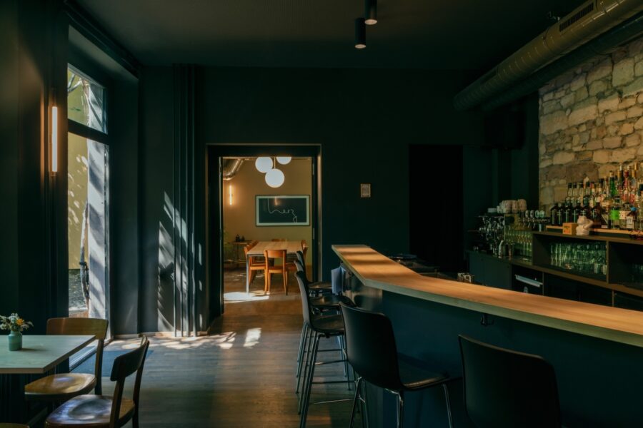 Modern dark bar interior with a long counter, stools, and a view into a sunlit dining room.