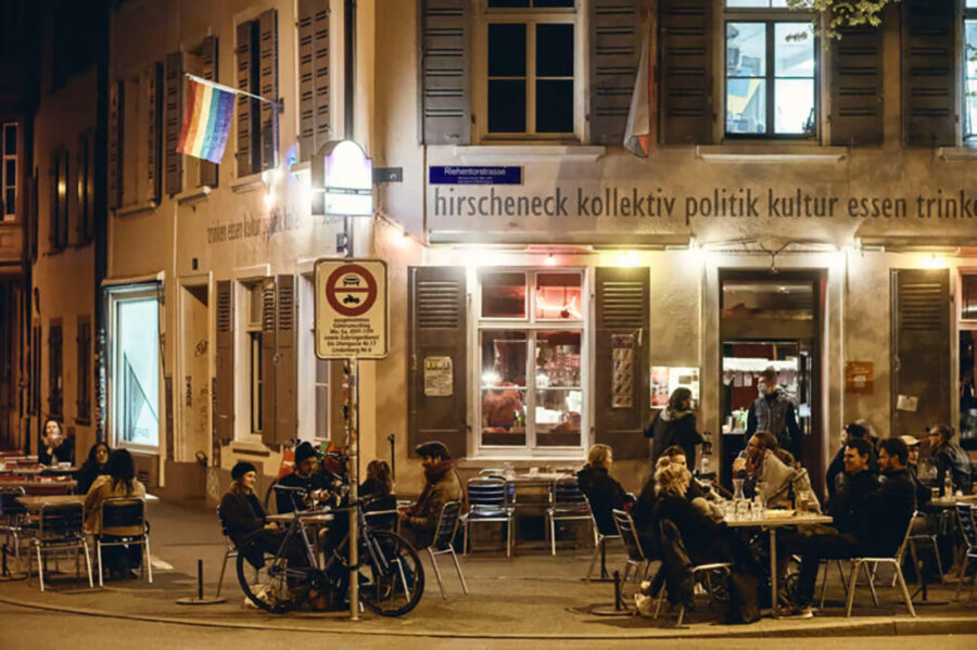 People sitting at outdoor cafe tables in front of a building with a rainbow flag.