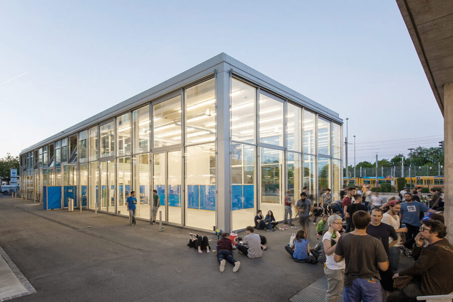 Ein modernes, hell erleuchtetes Glasgebäude mit Menschen, die davor stehen und auf dem Boden sitzen.