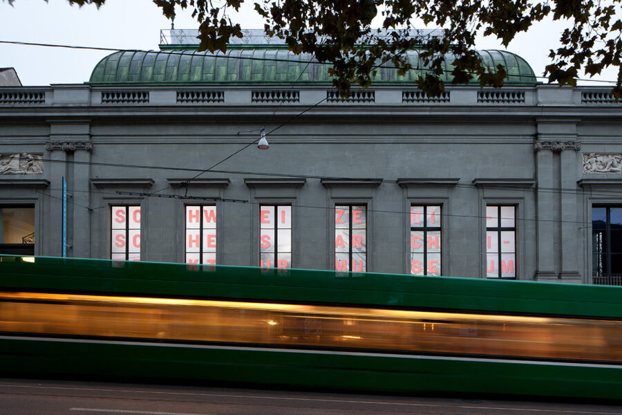 A blurred green tram passes a grey stone building with red lettering in the windows.