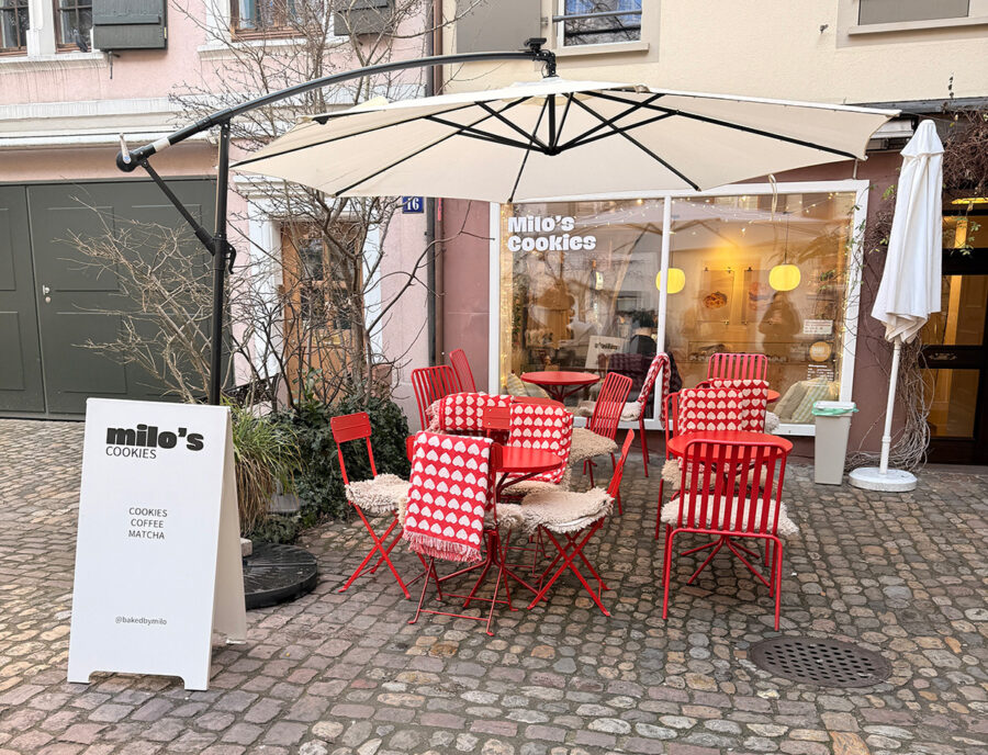 Outdoor seating area of Milo's Cookies with red chairs and tables under a large white umbrella.