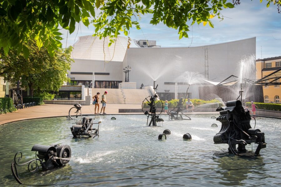 A fountain with mechanical sculptures spraying water in front of a modern white building.