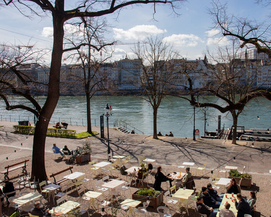 Outdoor cafe seating by a river with a city skyline and bare trees under a sunny sky.