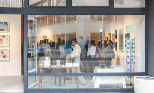 Blick in die Galerie während der Vernissage: Besucher:innen betrachten die ausgestellten Kunstwerke, im Vordergrund Tische mit Objekten und Katalogen.