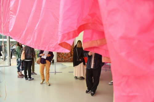 Menschen betrachten Kunstwerke in einer Galerie, teilweise verdeckt durch einen pinken Stoff.