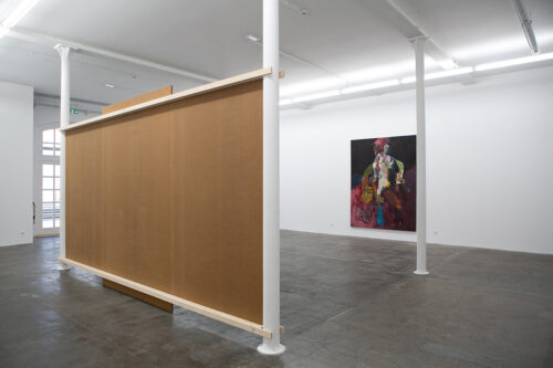 Heller Galerieraum mit einer großen braunen Trennwand und einem farbenfrohen Gemälde im Hintergrund.