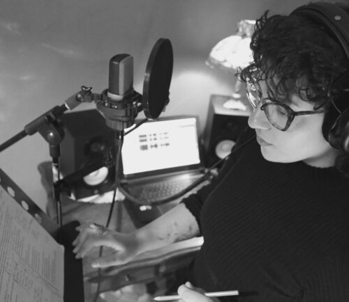 Schwarz-Weiß-Foto einer Person mit Kopfhörern und Brille vor einem Mikrofon in einem Studio.
