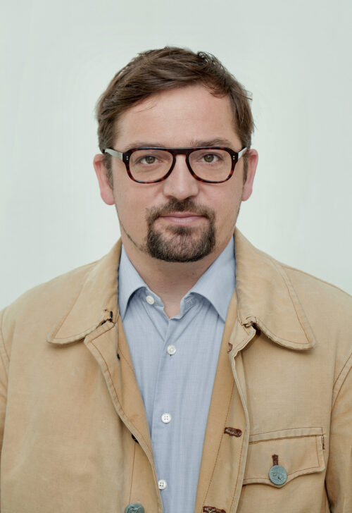 Porträt eines Mannes mit Brille, kurzem braunem Haar und Kinnbart in beige-blauer Kleidung.