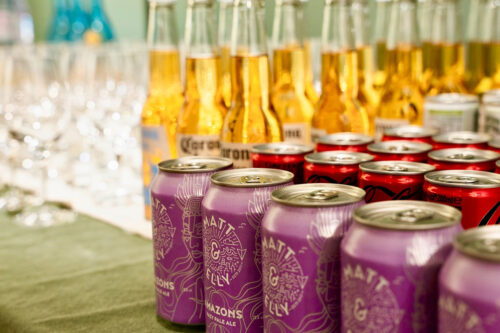 Lila Getränkedosen im Vordergrund, dahinter goldene Bierflaschen und Weingläser auf einem Tisch.