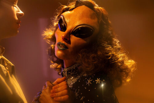 Nahaufnahme einer Person mit Alien-Maske, Lockenperücke und glitzerndem Kleid in warmem Licht.
