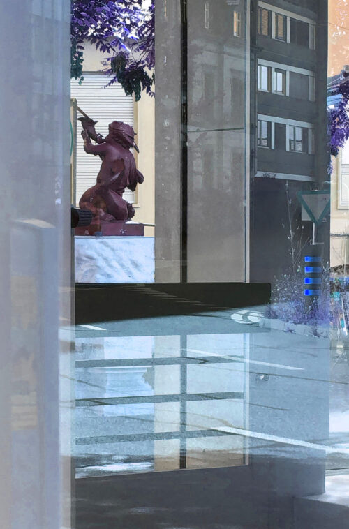 Spiegelung einer rötlichen Statue eines knienden Kriegers in einer Glasscheibe in der Stadt.
