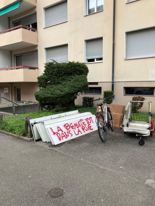Ein Fahrrad lehnt an weißen Fensterläden mit dem roten Graffiti-Schriftzug La Beauté est dans la Rue.