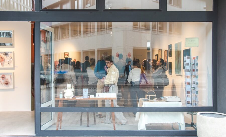 Blick in die Galerie während der Vernissage: Besucher:innen betrachten die ausgestellten Kunstwerke, im Vordergrund Tische mit Objekten und Katalogen.