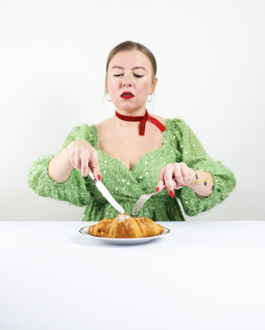 A woman in a green sequined dress uses a knife and fork to eat a croissant on a white table.