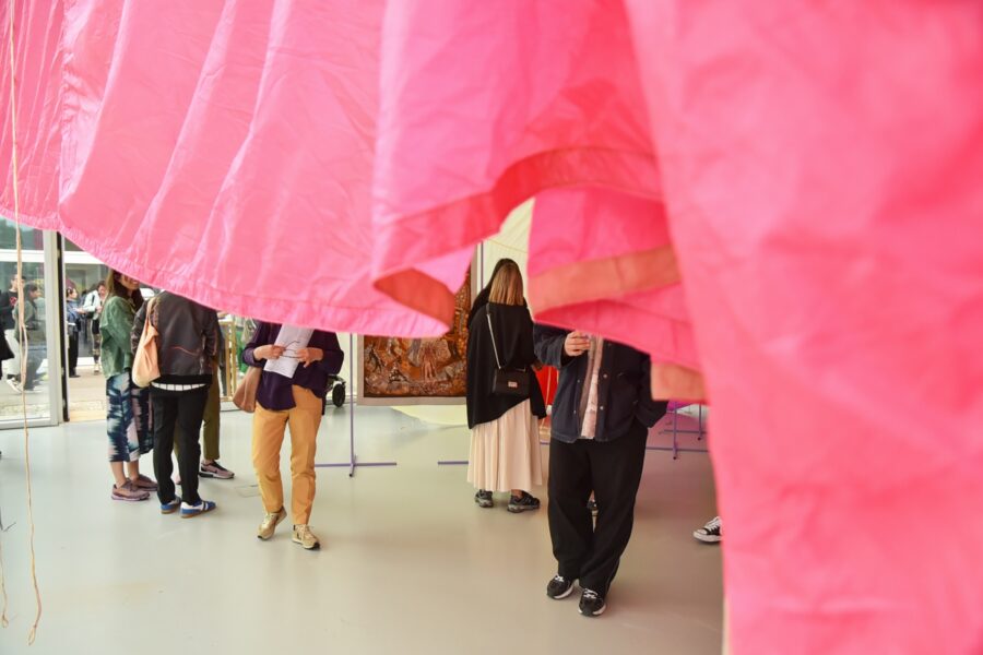 Menschen betrachten Kunstwerke in einer Galerie, teilweise verdeckt durch einen pinken Stoff.