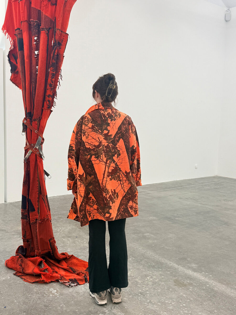 Frau von hinten in orange-braun gemusterter Jacke und schwarzer Hose neben einem roten Stoffbanner.