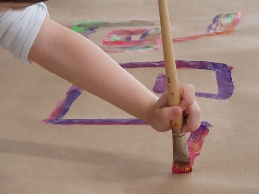 Ein Kind malt mit einem großen Pinsel bunte Formen auf braunes Papier.
