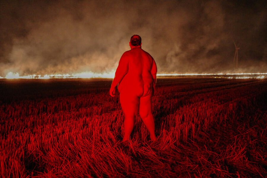 Nackter Mann von hinten in einem rot beleuchteten Getreidefeld vor einer Feuerlinie.