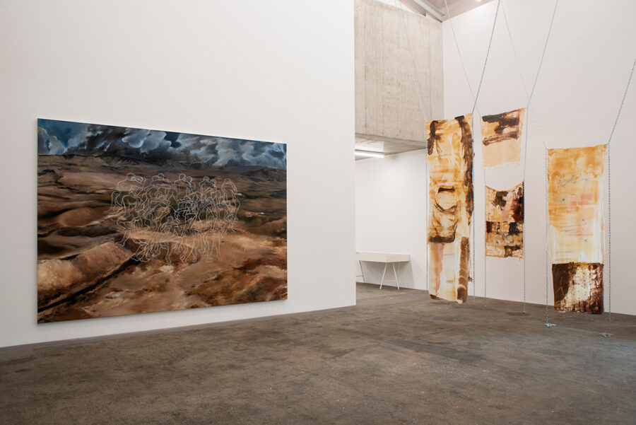 Ein Ausstellungsraum mit einem großen Wüstengemälde und hängenden, abstrakten Textilkunstwerken.