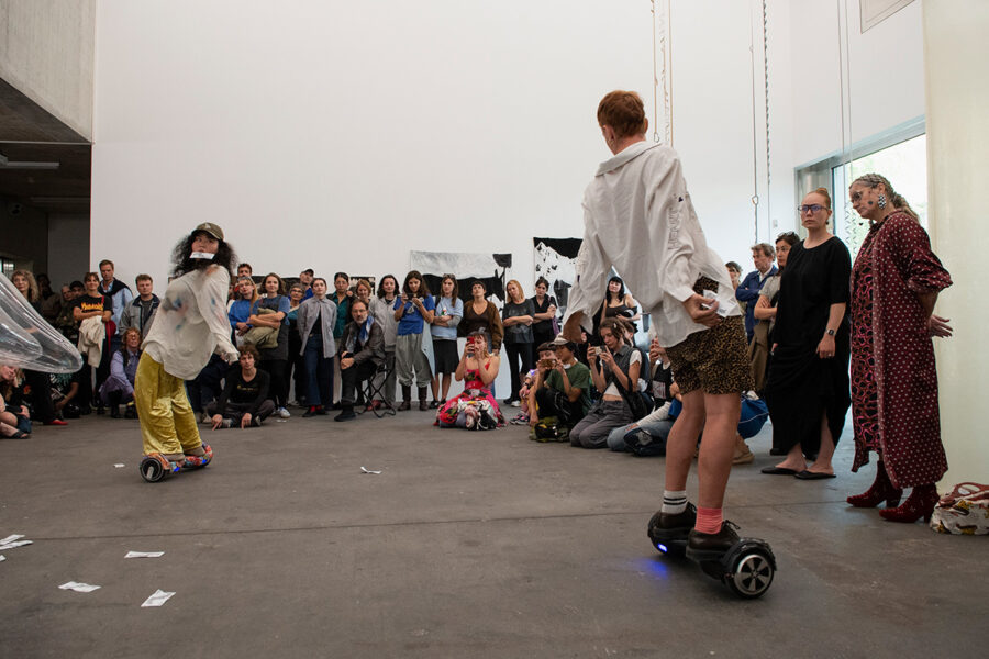 Zwei Personen auf Hoverboards performen in einem weißen Raum vor einem sitzenden Publikum.