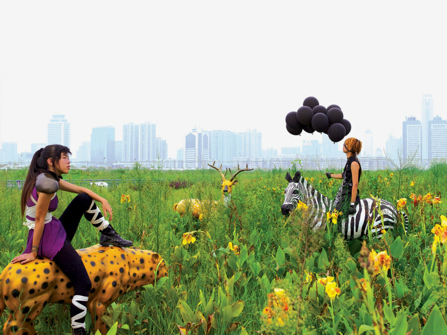 Two women in futuristic outfits sit on animal sculptures in a field with a city skyline backdrop.