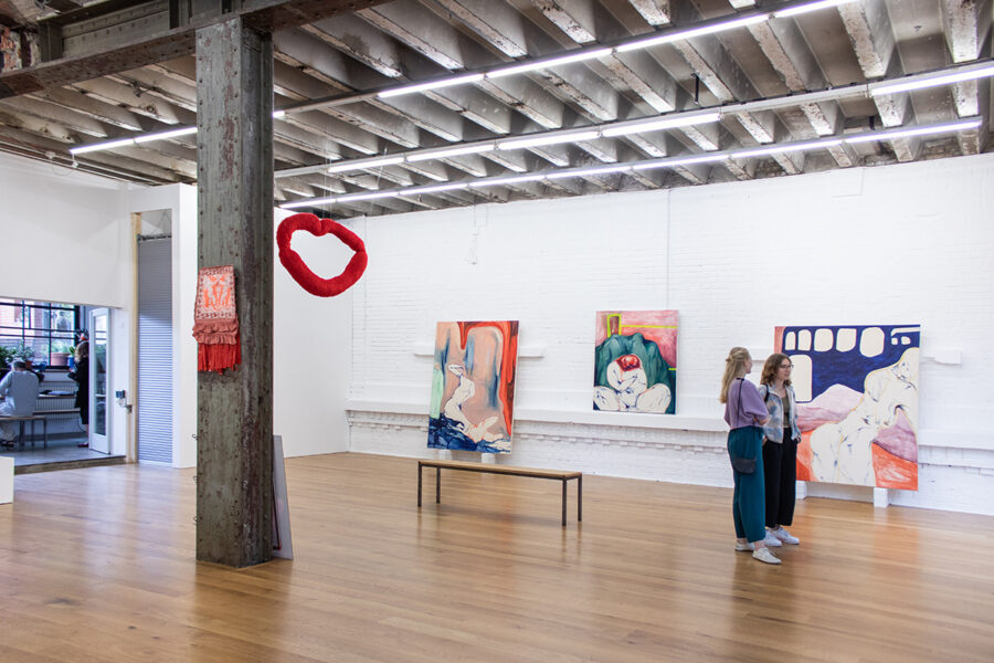 Two people view colorful abstract paintings in a bright, industrial art gallery.