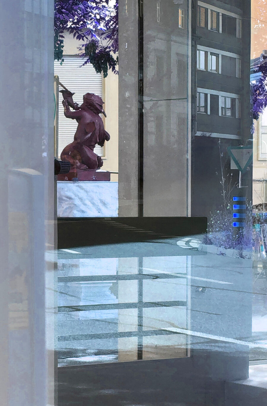 Spiegelung einer rötlichen Statue eines knienden Kriegers in einer Glasscheibe in der Stadt.