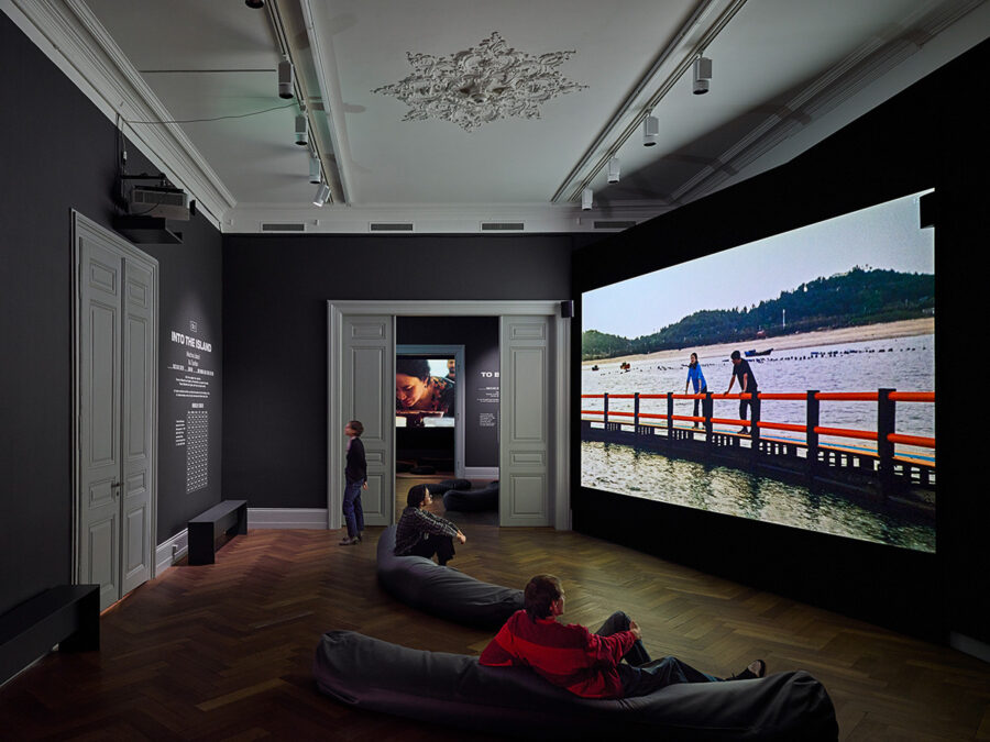People relaxing on beanbags in a dark gallery watching a large screen projection of a beach scene.