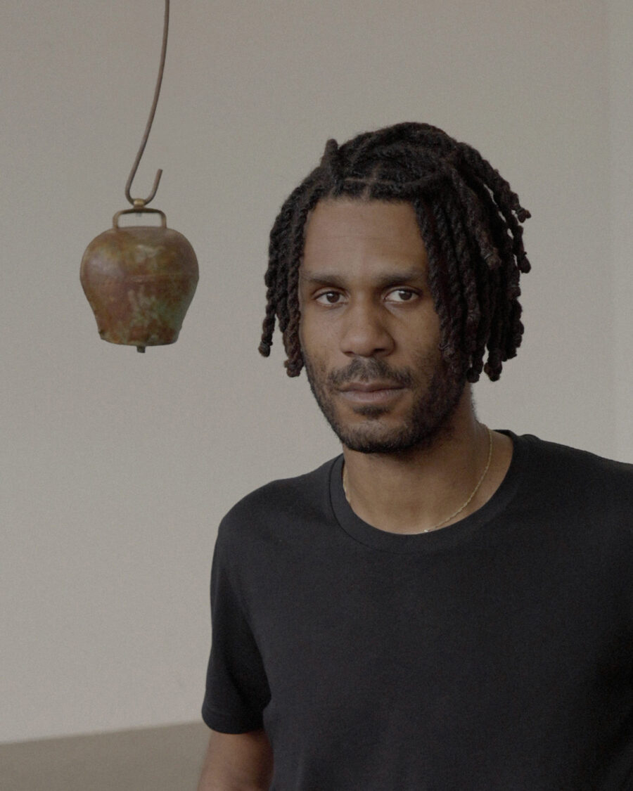 Portrait of a man with dreadlocks wearing a black t-shirt, with a hanging bell in the background.