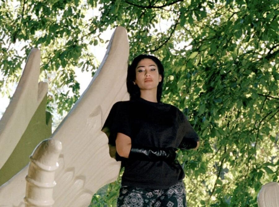 A woman in a black top and long gloves poses next to a white sculpture in a green forest.