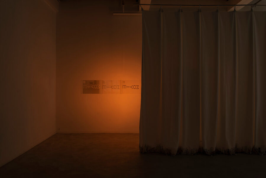 Ein dunkler Raum mit einem weißen Vorhang und einer orangefarbenen Lichtprojektion an der Wand.