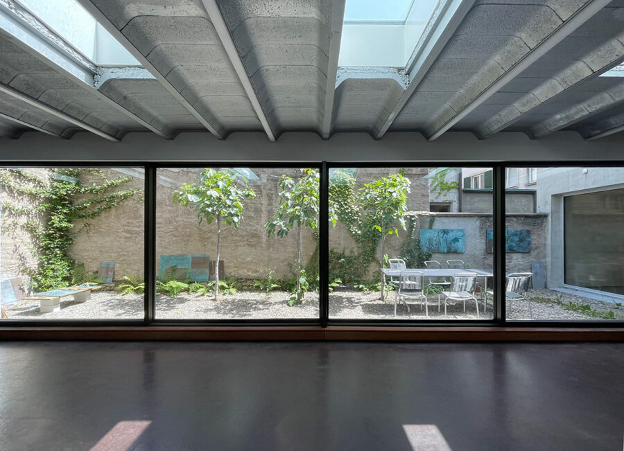 Modern indoor room with a concrete ceiling and large windows looking onto a green courtyard.