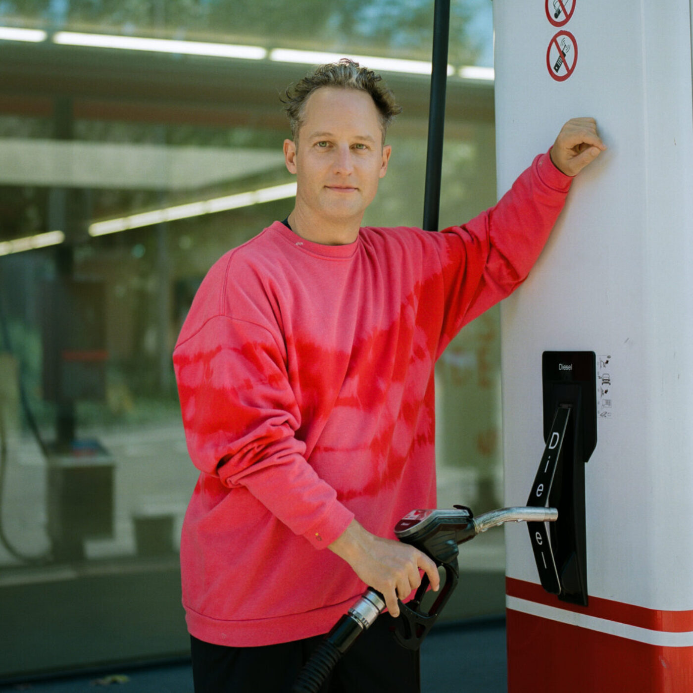 Ein Mann in einem rosa-roten Sweatshirt lehnt an einer Zapfsäule an einer Tankstelle.