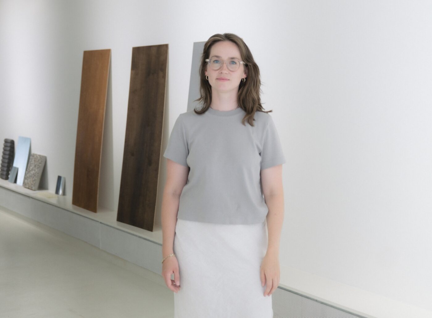 Eine Frau mit Brille, grauem T-Shirt und weißem Rock steht vor Holzpaneelen an einer weißen Wand.