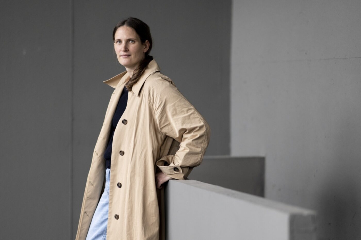 Frau in einem beigen Trenchcoat lehnt an einer grauen Betonmauer.
