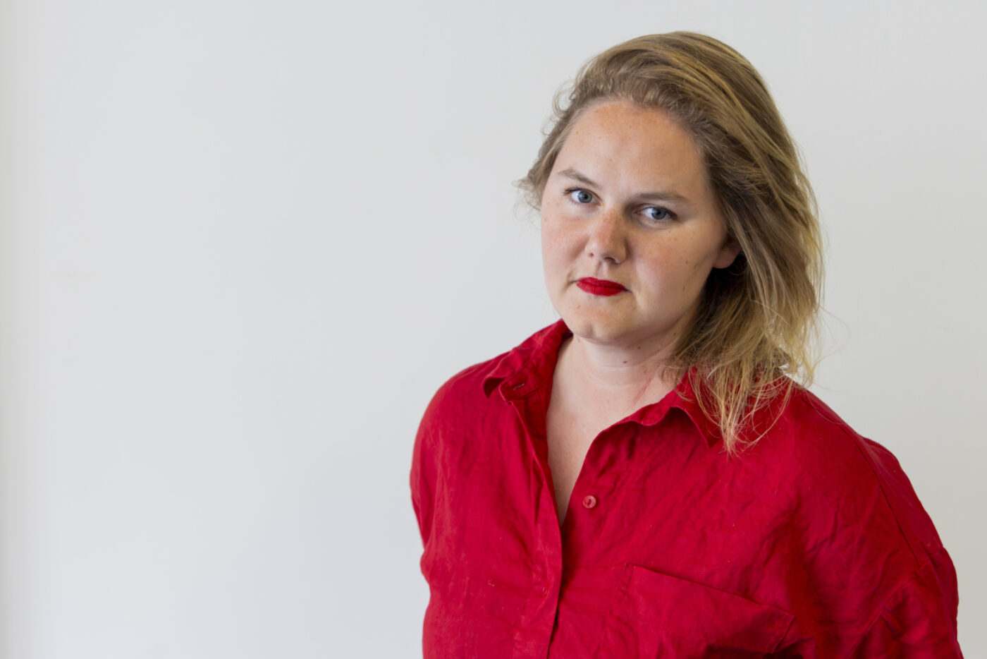 Porträt einer blonden Frau mit rotem Lippenstift und einer roten Bluse vor weißem Hintergrund.