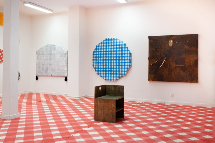 Kunstinstallation in einem weißen Raum mit kariertem Boden und verschiedenen Wandobjekten.