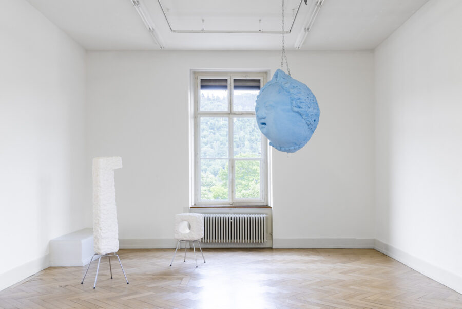 Ein heller Galerieraum mit einer hängenden blauen Kopfskulptur und weißen Kunstobjekten.