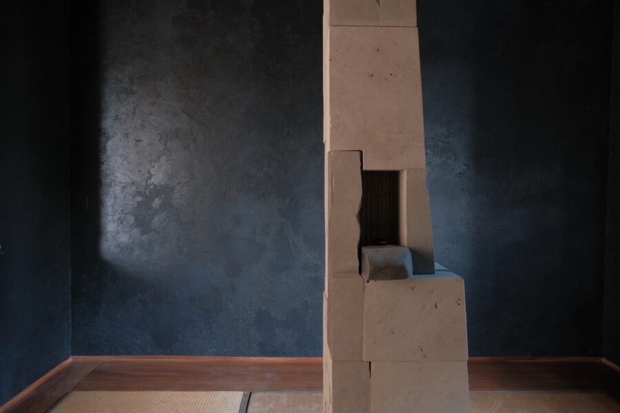 Ein minimalistischer, heller Steinkamin vor einer dunklen, strukturierten Wand in einem Tatami-Raum.