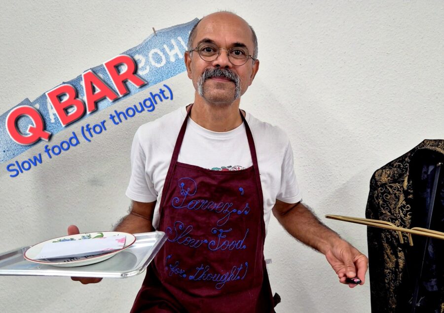 Ein Mann mit Brille und Schnurrbart trägt eine weinrote Schürze und hält ein Tablett.