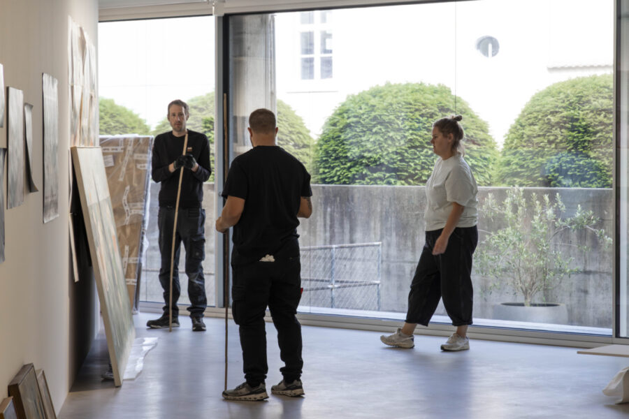 Drei Personen in Arbeitskleidung stehen in einem hellen Raum vor einer Wand mit Kunstwerken.