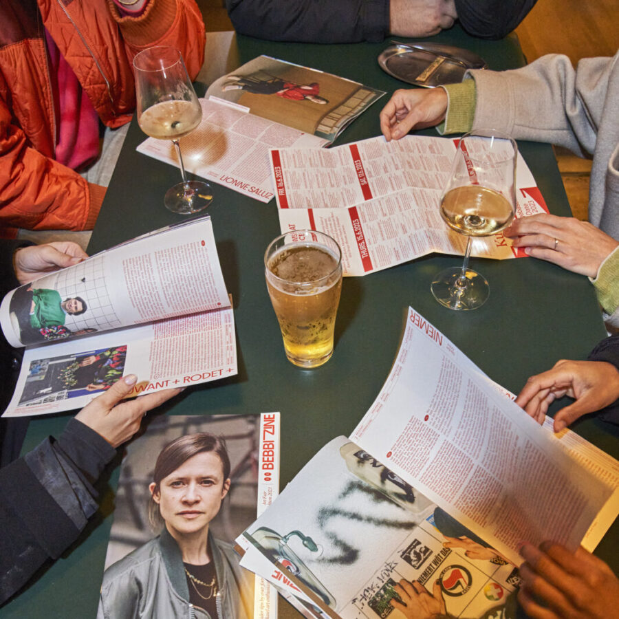 Menschen sitzen an einem Tisch mit Zeitschriften, Weingläsern und einem Bierglas.